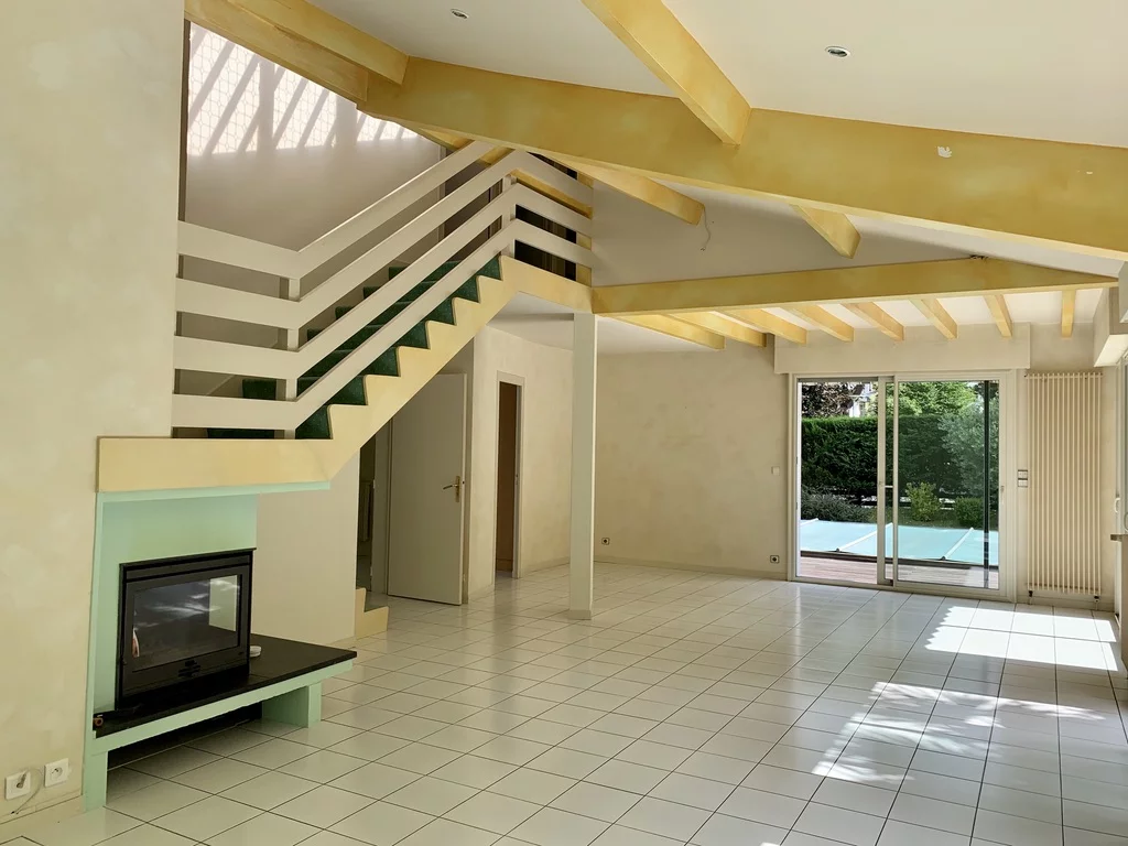 Photo avant rénovation d’un salon avec escalier design, cheminée, poutres apparentes et carrelage blanc, donnant sur une baie vitrée avec vue sur une piscine.